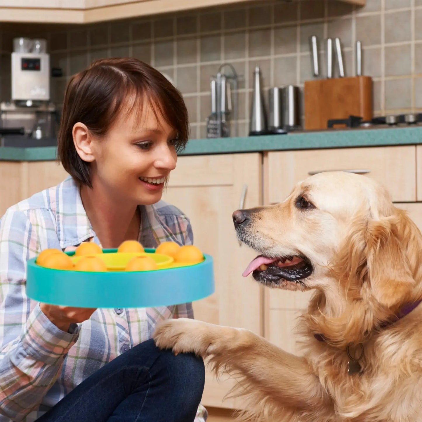 Woman holding a blue and green dog toy with a golden retriever in a kitchen.